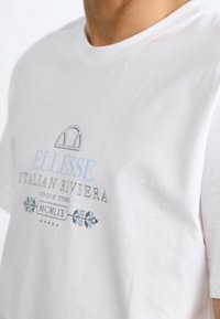 Valkoinen puuvillapaita, jossa on pyöreä kaula-aukko, jossa on sinisiä ja harmaita graafisia tekstejä, joissa lukee "ELLESSE ITALIAN RIVIERA", sekä koristeellisia kukkakuvioita.
