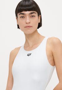 Λευκό μαγιό από λείο ύφασμα· διαθέτει στρογγυλή λαιμόκοψη και λεπτές τιράντες. Το λογότυπο «arena» είναι ορατό στο στήθος. Μινιμαλιστικός σχεδιασμός.