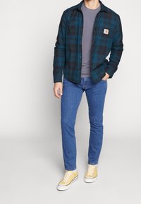 Rutig flanellskjorta i turkos och svart över en grå t-shirt, tillsammans med blå jeans och gula sneakers. Har knapppå detaljer på skjortan.