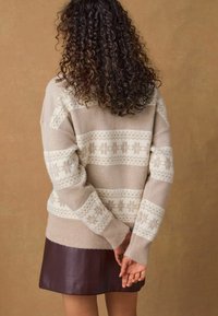 Pullover beige a maglia con motivi geometrici bianchi, taglio oversize, polsini a coste, abbinato a una gonna in pelle bordeaux. Sfondo marrone uniforme.