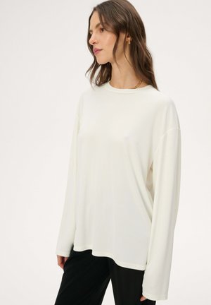 Kobieta ubrana w luźną, białą bluzkę z długim rękawem i czarne spodnie, zwrócona w lewo, z minimalistyczną biżuterią na jednolitym tle.