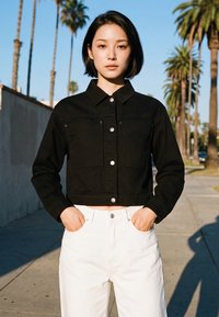 Fiatal nő rövid sötét hajjal napsütötte járdán áll, pálmafák között, fekete gombos dzsekit és fehér nadrágot visel, zsebében tartja a kezét.