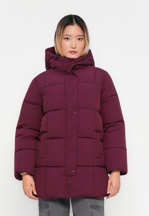 Femme portant une doudoune d'hiver violet foncé avec capuche, boutons-pression à l'avant et manches longues, se tenant devant un fond uni.
