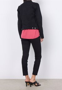 Chaqueta de denim negra con detalles de botones, llevada sobre una blusa rosa. Pantalones negros ajustados y sandalias de tacón negro completan el conjunto.