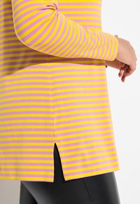 Maglietta a maniche lunghe a righe in rosa e giallo. Presenta una linea dritta con spacchi laterali e un tessuto morbido e leggero, abbinata a leggings neri.