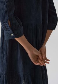 Vestito blu navy con maniche a tre quarti, caratterizzato da un dettaglio a bottoni sul polsino e un design a gonne a strati. Mani giunte dietro la schiena.