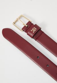 Ceinture en cuir rouge avec boucle en métal doré. Présente un logo en relief près de la boucle et des trous ronds pour les ajustements le long de la sangle.
