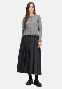 Grau gestrickte Wolle Cardigan mit durchgehender Knopfleiste und Rüschensaum, kombiniert mit einem langen schwarzen Rock. Das Modell trägt schwarze Schuhe und Socken.