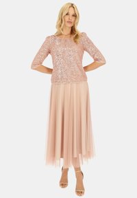 Różowa bluzka z cekinami i rękawami 3/4, noszona na zwiewnej, sięgającej kostek spódnicy z tiulu w pasującym, jasnoróżowym odcieniu. Szpilki z paskami wokół kostki.