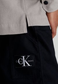 Jasnoszara koszula o fakturze z detalem w postaci guzika, zestawiona z ciemnonawigowymi spodniami, które mają czarną prostokątną naszywkę z napisem "Calvin Klein Jeans."