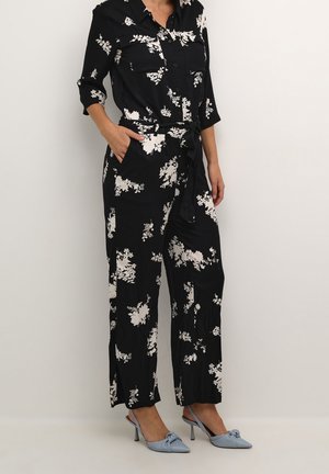 Schwarzer Jumpsuit mit floralen und weißen Mustern, Knopfleiste vorne, zwei Brusttaschen, gebundenem Taillenband und weit geschnittenen Hosenbeinen. Kombiniert mit hellblauen Absätzen.