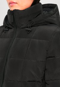 Nærbilde av person som har på seg svart, quiltet Armani Exchange-jakke med høy krage og hette, delvis glidd opp.