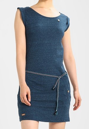 Vrouw draagt een mouwloze blauwe jurk met een gevlochten riem gestrikt in de taille en zakken aan beide zijden.