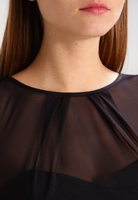 Schwarze transparente Bluse mit Rundhalsausschnitt, dezenten Falten am Halsauschnitt und einer Schicht aus festem schwarzem Stoff darunter.