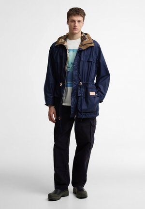 Młody mężczyzna w granatowej kurtce z kapturem i ściągaczem w talii, z grafiką na koszulce, czarnych spodniach bojówkach oraz zielonych wsuwanych butach, stojący przodem.