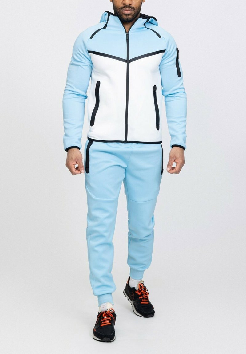 Survêtement de sport bleu clair avec un devant blanc, featuring des accents noirs, des poches zippées et une capuche. Design texturé et ajusté.