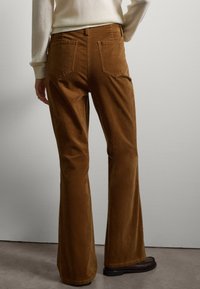 Braune Corduroy Schlaghosen mit zwei Gesäßtaschen und genähten Details, kombiniert mit einem hellen Pullover und braunen Loafers.