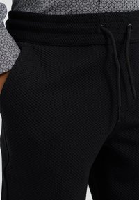 Joggers neri in tessuto strutturato con vita elasticizzata e cordoncino. Primo piano che mostra un sottile motivo a waffle e una camicia chiara a contrasto.