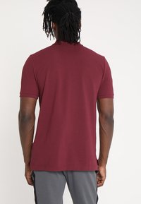 Bordeauxfarbenes Poloshirt mit kurzen Ärmeln, geripptem Kragen und glatter Textur. Taillierte Passform mit geradem Saum. Etikett am unteren Ende sichtbar.