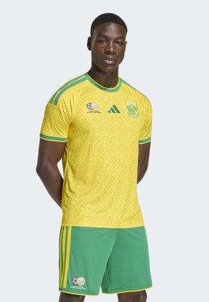 Mužský atlét oblečený v žlto-zelenom drese a šortkách juhoafrického futbalového tímu, stojaci s rukami za chrbtom na jednoduchom pozadí.