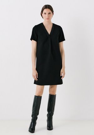 Vestito nero a maniche corte con scollo a V, caratterizzato da cuciture decorative lungo il collo. Abbinato a stivali neri al ginocchio.