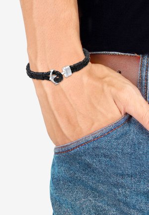 Sort flettet læderarmbånd med sølvfarvet lås og dekorativ knude detalje, båret på et håndled, mod denimstof.