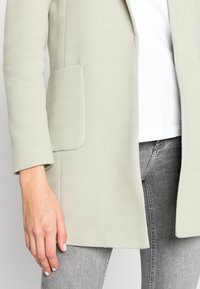 Jasnozielony płaszcz w jodełkę z klapami i boczną kieszenią, w zestawieniu z białą koszulą i szarymi dżinsami. Gładka tekstura, dopasowany krój.