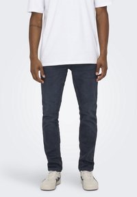 Jeans azul oscuro ajustados de denim, con cinco bolsillos y una textura suave, combinados con una camiseta blanca y zapatillas ligeras.