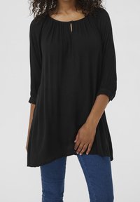 Tunique noire avec un col rond, détail en trou de serrure et texture froncée. Dispose de manches trois-quarts avec poignet. Associée à un jean bleu.