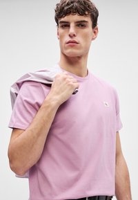 Lys rosa bomull t-skjorte med rund hals og korte ermer, med et lite logo patch på brystet. Modellen holder en stripete skjorte.