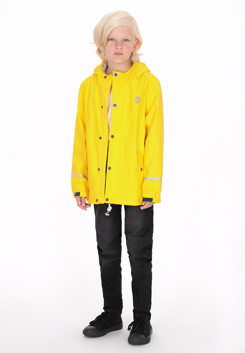 Gul vattentät jacka med huva, tryckknappar, reflexdetaljer, bärs över svarta slim fit jeans och svarta sneakers.