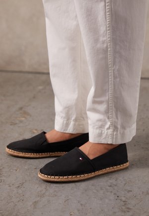 Person iført hvide bukser og sorte espadrilles med flettede såler, stående på en beton gulv.