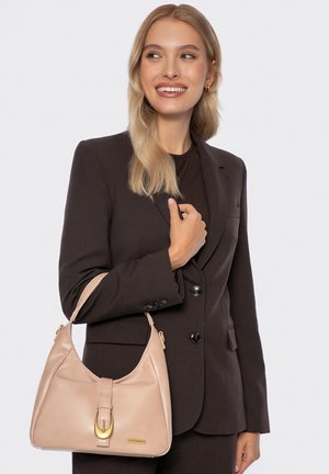 Femme en costume marron foncé tenant un sac à main en cuir beige avec une boucle dorée, souriante et regardant sur le côté devant un fond uni.