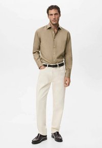 Camisa color marrón claro con botones, pantalones crema y cinturón negro. Mocasines negros pulidos, en pose de pie frente a un fondo blanco liso.