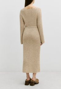 Beige, gestricktes langes Kleid mit einem strukturierten Muster und langen Ärmeln. Hat eine taillierte Taille mit einem passenden Gürtel. Chunky Heeled Mules sind sichtbar.
