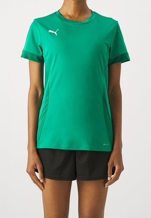 Persoon die een groen Puma sportshirt met korte mouwen en zwarte shorts draagt, staande tegen een effen lichte achtergrond.