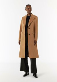 Langer, maßgeschneiderter Mantel aus einem camel-farbenen Wollmischgewebe mit Schalkragen, zwei Knöpfen und seitlichen Taschen, über einem schwarzen Oberteil und einer schwarzen Hose, kombiniert mit schwarzen Stiefeln.