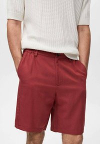 Shorts rouges en coton avec poches, présentant un devant plat et un bouton foncé contrastant. Portés avec une chemise en tricot blanche texturée.