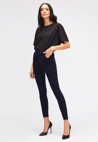 Chemise noire à manches courtes associée à un jean skinny taille haute bleu foncé et des escarpins noirs à bout pointu. Design simple et ajusté.