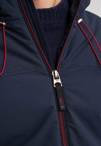 Jachetă navy cu o textură netedă, având accente roșii la fermoar, șnururi elastice pe glugă și căptușeală din tricot gri.