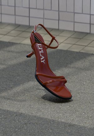 Red strappy kitten heel sandal with a triangular silver buckle and "REPLAY" branding on the insole, on a tiled and asphalt floor.