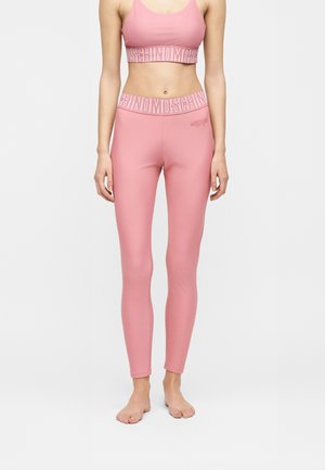 Vrouw draagt een roze Moschino sportswear set met sportbeha en legging, beide met witte Moschino logo bandjes in de tailleband.