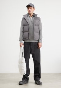 Chaleco acolchado gris sobre un suéter con cremallera gris claro, pantalones negros y botas negras gruesas, sosteniendo una bolsa blanca mullida.
