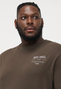 Brun bomulds T-shirt med korte ærmer, med hvid trykt tekst i en stiliseret skrifttype på venstre brystområde.