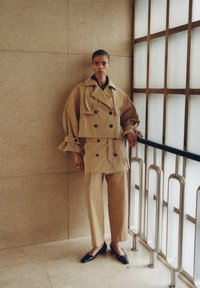 Trench-coat beige à double boutonnage avec des manches surdimensionnées, associé à un pantalon tailleur assorti et des ballerines noires. Tissu lisse, détails minimalistes.