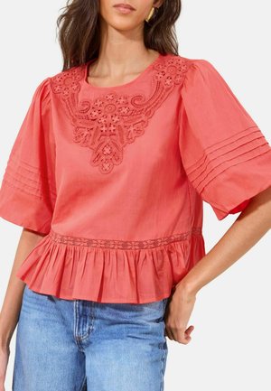 Žena v koralovej blúzke s kvetinovou čipkou na ramenách, záhybkovými rukávmi, plisovaným okrajom a svetlomodrými džínami na bielom pozadí.