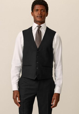 Gilet noir avec un décolleté en V, fermeture à cinq boutons et deux poches avant. Porté sur une chemise blanche et une cravate à motifs.