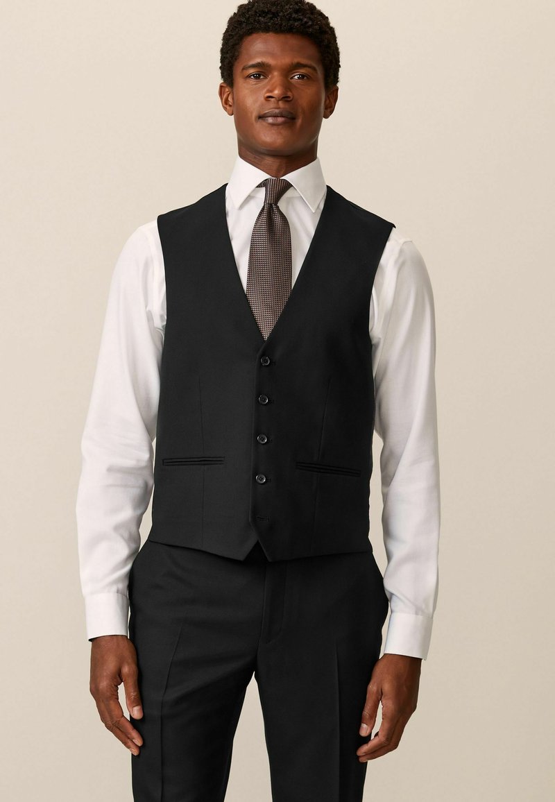 Chaleco negro con un diseño de escote en v, cierre de cinco botones y dos bolsillos frontales. Llevado sobre una camisa blanca y una corbata con estampado.