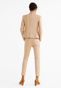 Beżowy garnitur szyty na miarę z dopasowaną marynarką i wąskimi spodniami. Tkanina wydaje się gładka, o czystym, minimalistycznym wzornictwie. Pasujące brązowe szpilki.