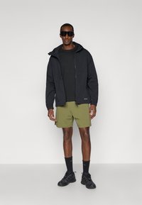 Veste à capuche noire sur une chemise noire, associée à un short vert olive. Les caractéristiques incluent des lunettes de soleil sombres et des chaussures de sport noires avec des chaussettes.